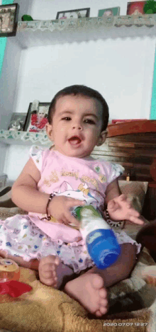 a baby girl in a pink dress is sitting on a bed holding a blue bottle .