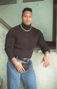 a man wearing a black turtleneck and blue jeans is leaning on a wall