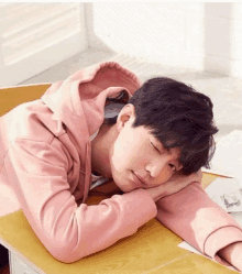 a young man in a pink hoodie is sleeping on a wooden desk