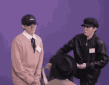 two men are standing next to each other on a purple background . one of the men is wearing a hat .