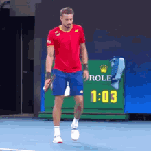 a man in a red shirt and blue shorts is walking on a tennis court with a rolex sign behind him