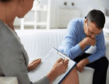 a man is sitting on a couch talking to a woman who is holding a clipboard and writing on it .