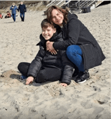 a woman is hugging a boy who is sitting in the sand on the beach