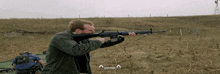 a man in a green jacket is holding a rifle in a field with the letters a and a below him