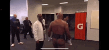 a man without a shirt is standing in a locker room next to a man in a white jacket .