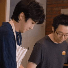 two men are standing next to each other in a kitchen preparing food . one of the men is wearing glasses and an apron .