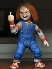 a chucky doll is giving the middle finger while wearing blue overalls