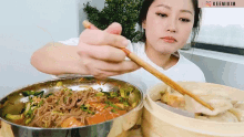 a woman in a white shirt is eating noodles with chopsticks from a bowl with the name keemikim on the bottom right