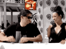 a man and a woman are sitting at a table in front of a wall with wb logos on it .