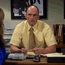 a man in a yellow shirt and tie is sitting at a desk in front of a poster that says mm