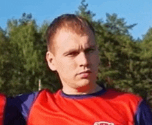 a man in a red and blue shirt is standing in front of trees and looking at the camera .