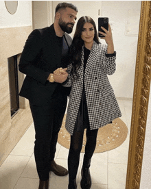 a man in a suit and a woman in a coat are taking a selfie in front of a mirror