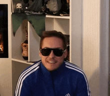 a man wearing sunglasses and an adidas jacket smiles