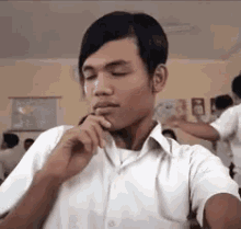 a young man in a white shirt is sitting in a classroom with his eyes closed and his hand on his chin .