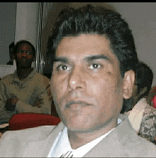 a man in a suit and tie is sitting in front of a group of people and looking at the camera .