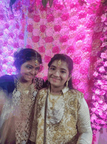 two girls pose for a picture in front of a wall of flowers