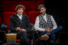 two men sit next to each other in a theater with a popcorn bucket in front of them