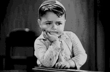 a black and white photo of a little boy sitting at a desk with his hand on his chin .