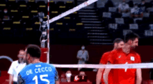 a volleyball player wearing a blue jersey with the number 15 on it is standing in front of a volleyball net .