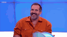 a man with a beard is laughing with his arms crossed in front of a blue background with the bbc logo on it