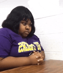 a woman in a purple shirt is sitting at a table with her hands folded in front of her .