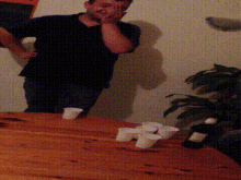 a man in a black shirt is covering his face while standing in front of a table with cups and a bottle on it