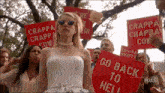 a woman in a white dress stands in front of a crowd holding signs that say go back to hell