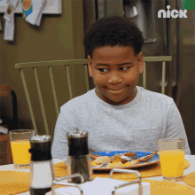 a young boy sits at a table with a plate of food and a glass of orange juice with a nick logo behind him