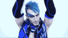 a man with blue hair and black elbow pads is standing with his arms in the air and looking at the camera .