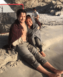 a man and a woman wrapped in blankets are sitting on the beach