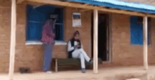 a woman is sitting on a bench on the porch of a house while a man stands in the window .