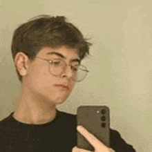 a young man wearing glasses is taking a selfie with his cell phone in front of a mirror .