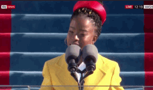 a woman wearing a yellow jacket and red hat stands in front of two microphones with sky news in the corner