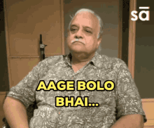 a man with a mustache is sitting in front of a sign that says " aage bolo bhai "