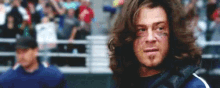 a man with long hair and a beard is standing in front of a crowd at a baseball game .