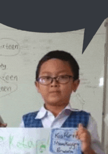 a young boy wearing glasses and a blue vest is holding a sign in front of a whiteboard .