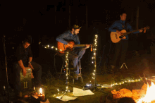 three men playing guitars in front of a fire with one wearing a hat that says ' a ' on it