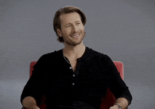 a man in a black shirt is sitting in a red chair smiling