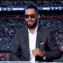 a man wearing sunglasses and a suit is standing in front of a stadium that says costa rica on the scoreboard