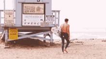 a man carrying a surfboard walks towards a lifeguard tower with a sign that says " caution beware of "