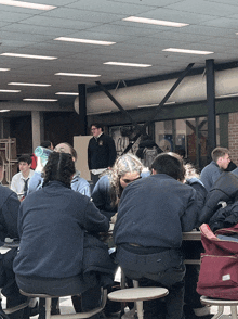 a group of people sit at a table in a cafeteria with a man standing behind them