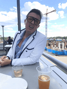 a man wearing sunglasses and a white jacket sits at a table with a cup of beer