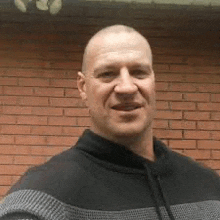 a bald man in a black and gray sweater is standing in front of a brick wall .