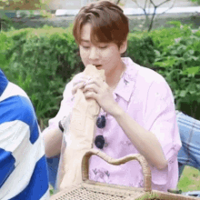 a young man in a pink shirt is eating a sandwich from a bag while sitting in a park .