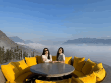 two women sit on a couch with yellow pillows in front of mountains