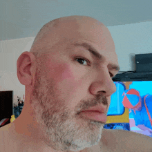 a bald man with a beard looks at the camera in front of a television