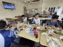 a group of men are sitting at tables eating food with a coca cola fridge in the background