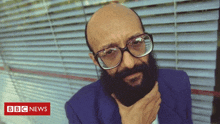 a bald man with glasses and a beard is standing in front of a bbc news sign