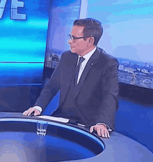 a man in a suit and tie stands at a news desk with a glass of water in front of him