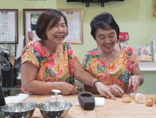 two women are laughing while preparing food in front of a wall with a picture of a man on it